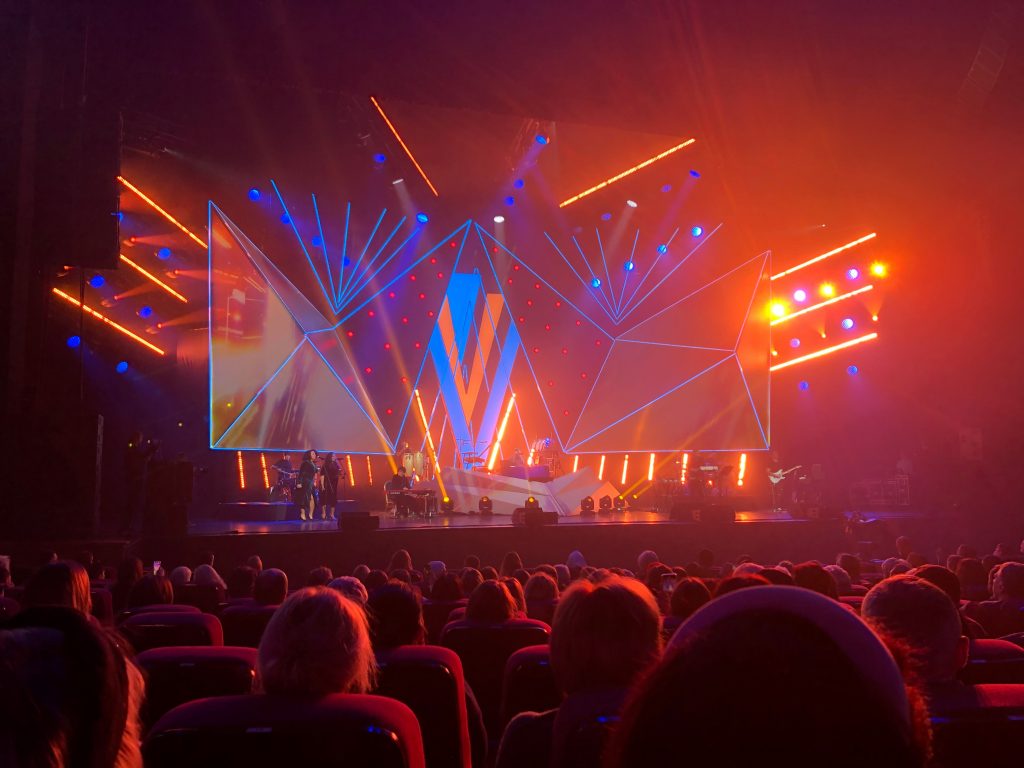 Escenario de concierto con pantallas LED laterales mostrando al artista en directo, iluminación escénica profesional y público asistiendo al espectáculo