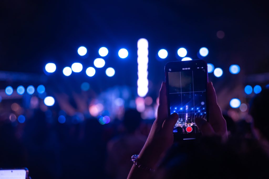 Mano grabando en vídeo un concierto nocturno con smartphone, luces de escenario desenfocadas en azul al fondo